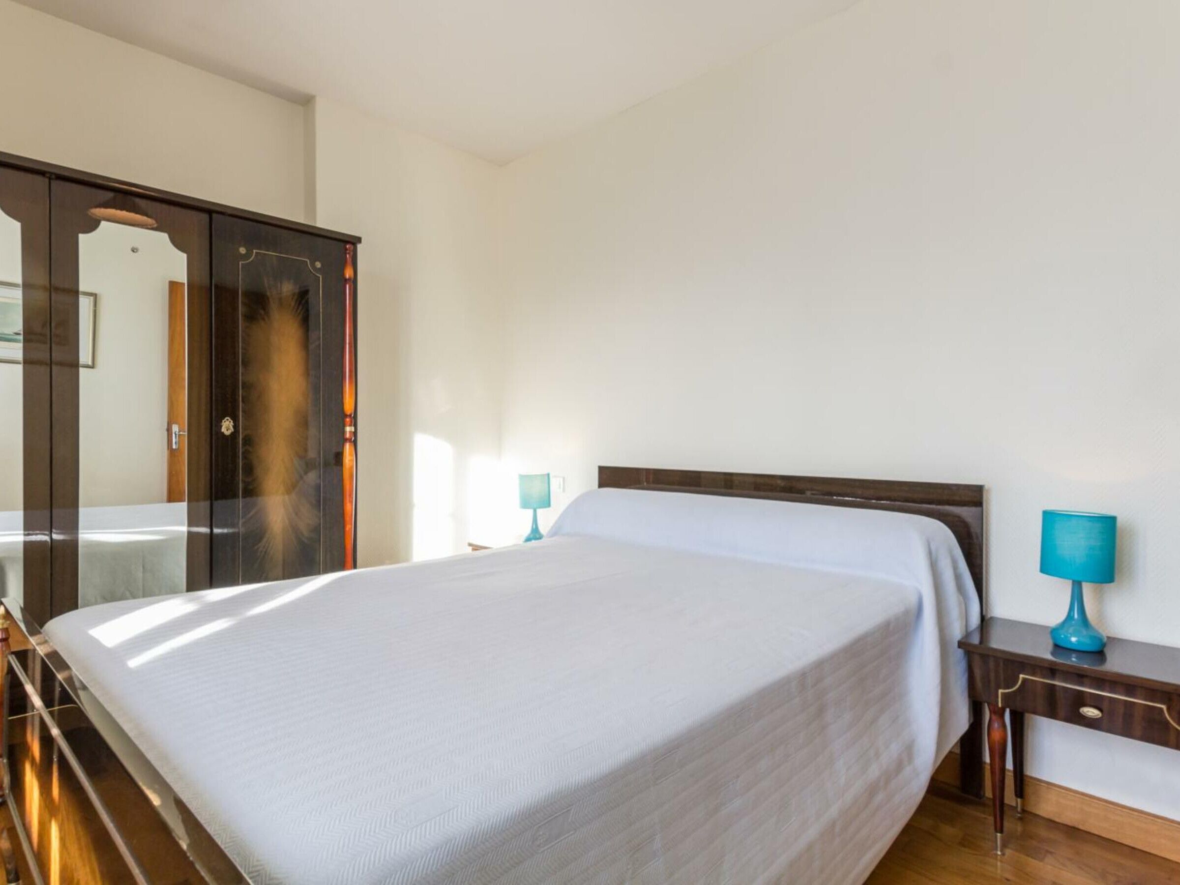 A neatly made bed in a room with a wooden wardrobe and a nightstand with a lamp.
