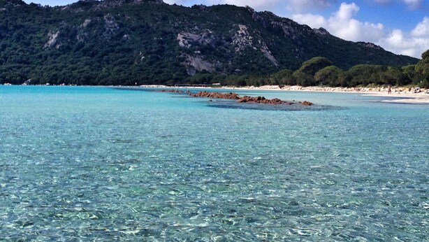 Een strand met helder turquoise water, rotsachtige rotsformaties en een bebost gebergte op de achtergrond.