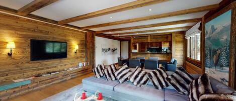 A cozy living room with a wooden interior, a flat-screen TV mounted on the wall, a fireplace, and a dining area with a kitchen in the background.