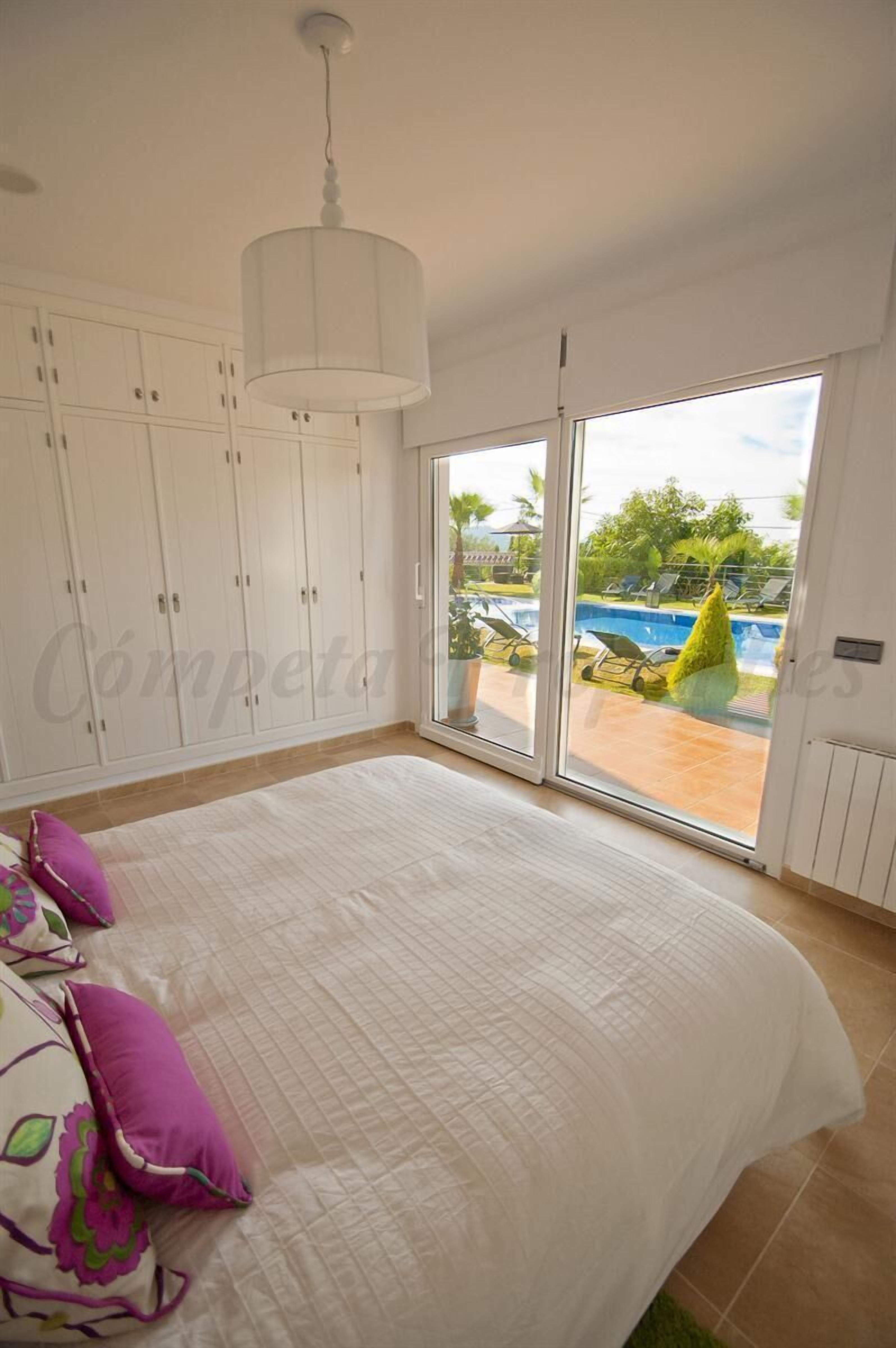 A bedroom with a large bed, white curtains, and a view of a swimming pool.