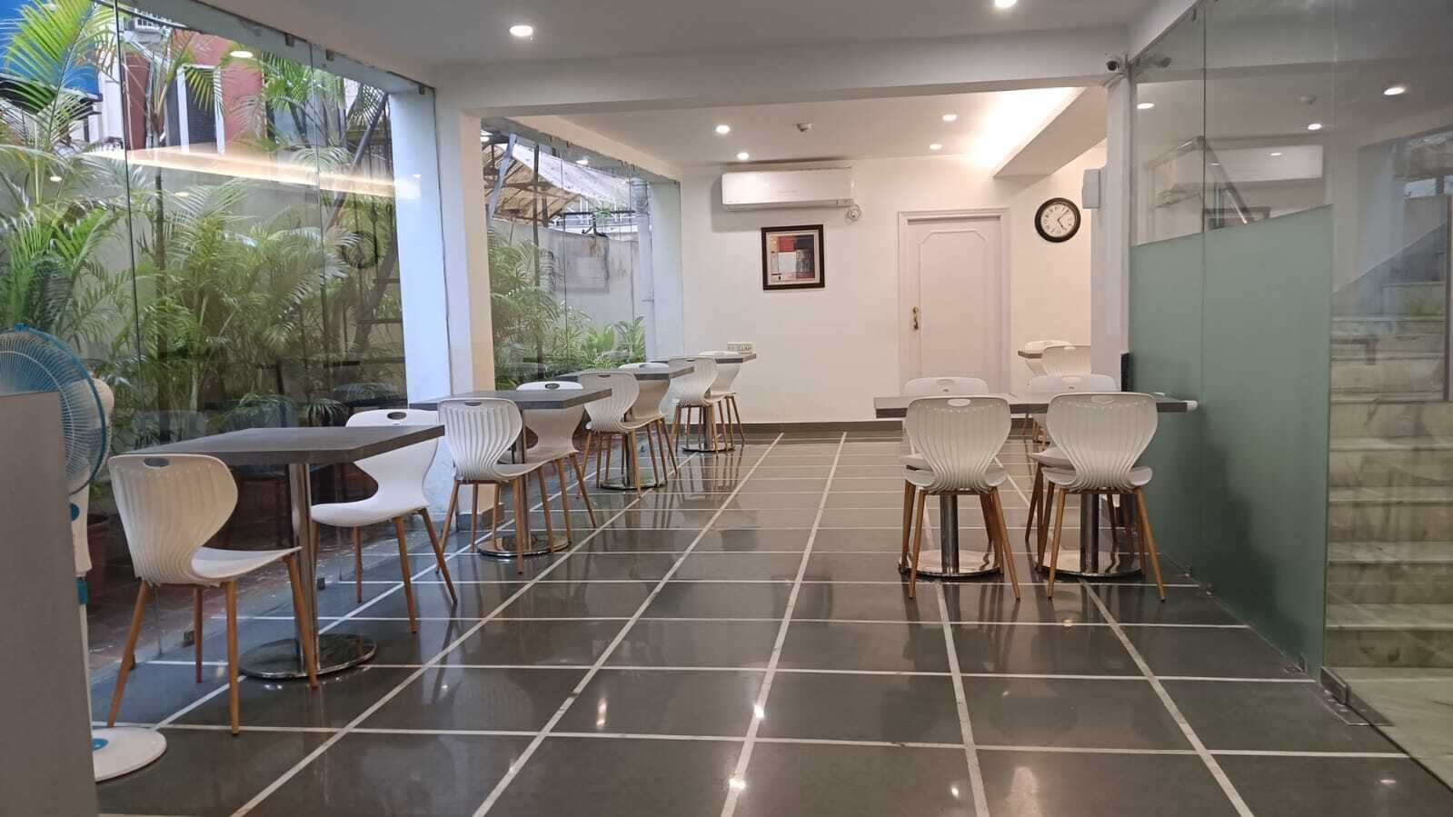 Un espace repas moderne avec une longue table et des chaises, une horloge au mur et une vue sur la verdure grâce à de grandes fenêtres.