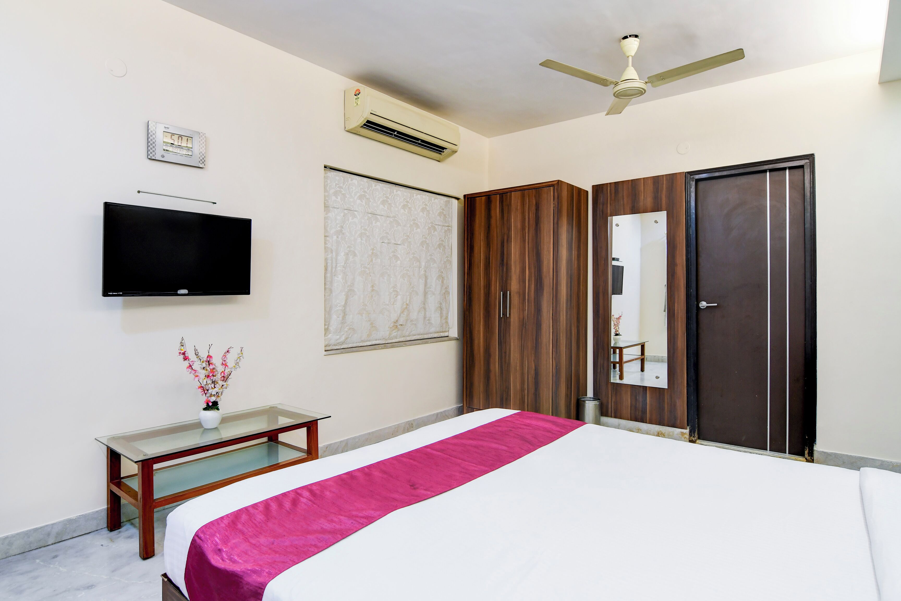 A hotel room with a bed, a flat-screen TV, a ceiling fan, an air conditioning unit, a wooden wardrobe, and a small table with a vase of flowers.
