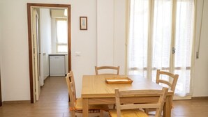 Comedor con mesa y sillas de madera, cocina al fondo y vista a través de una ventana con cortinas.
