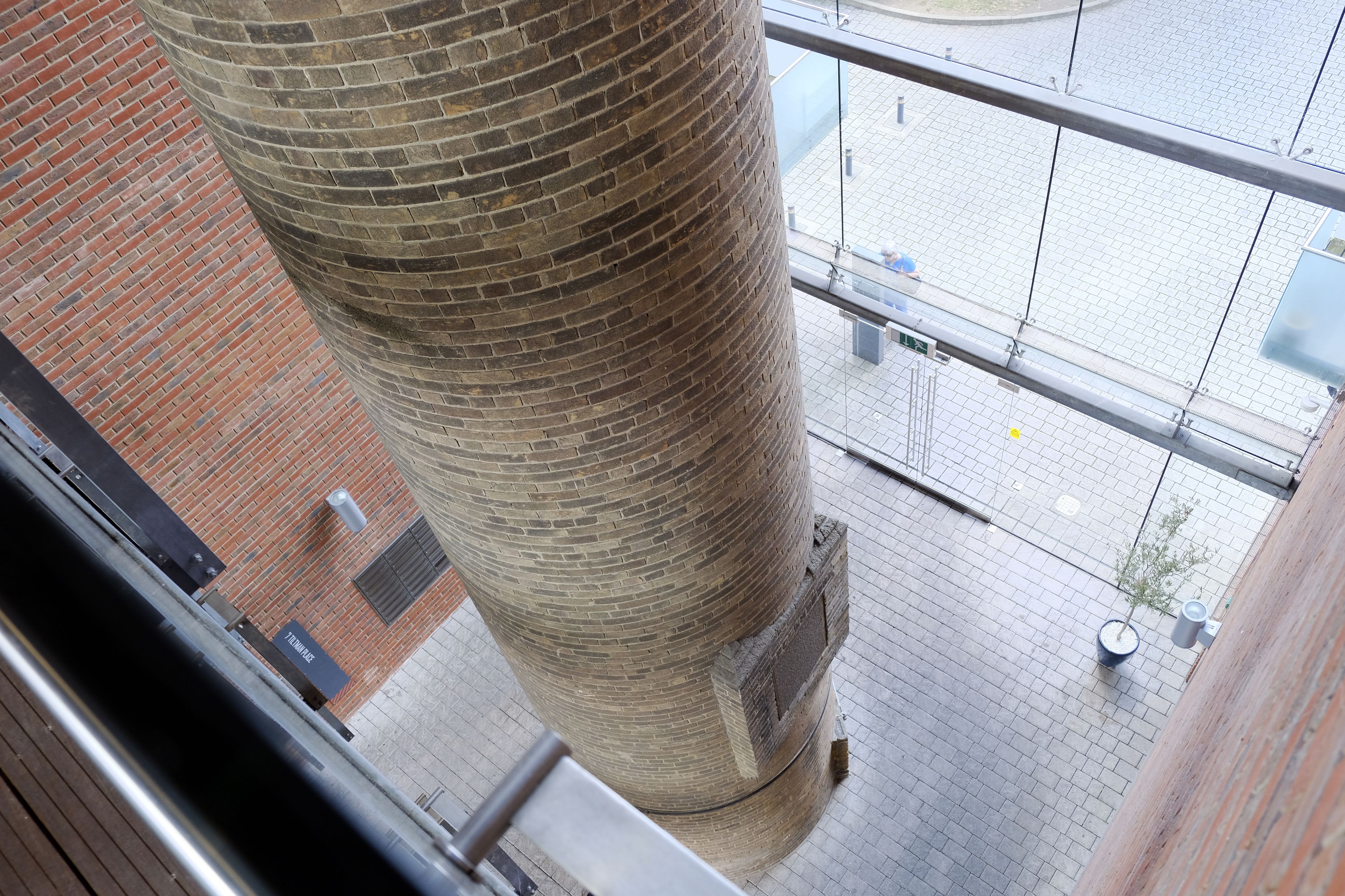 A view of a tall brick column from a balcony, with a brick building and a tiled plaza below.