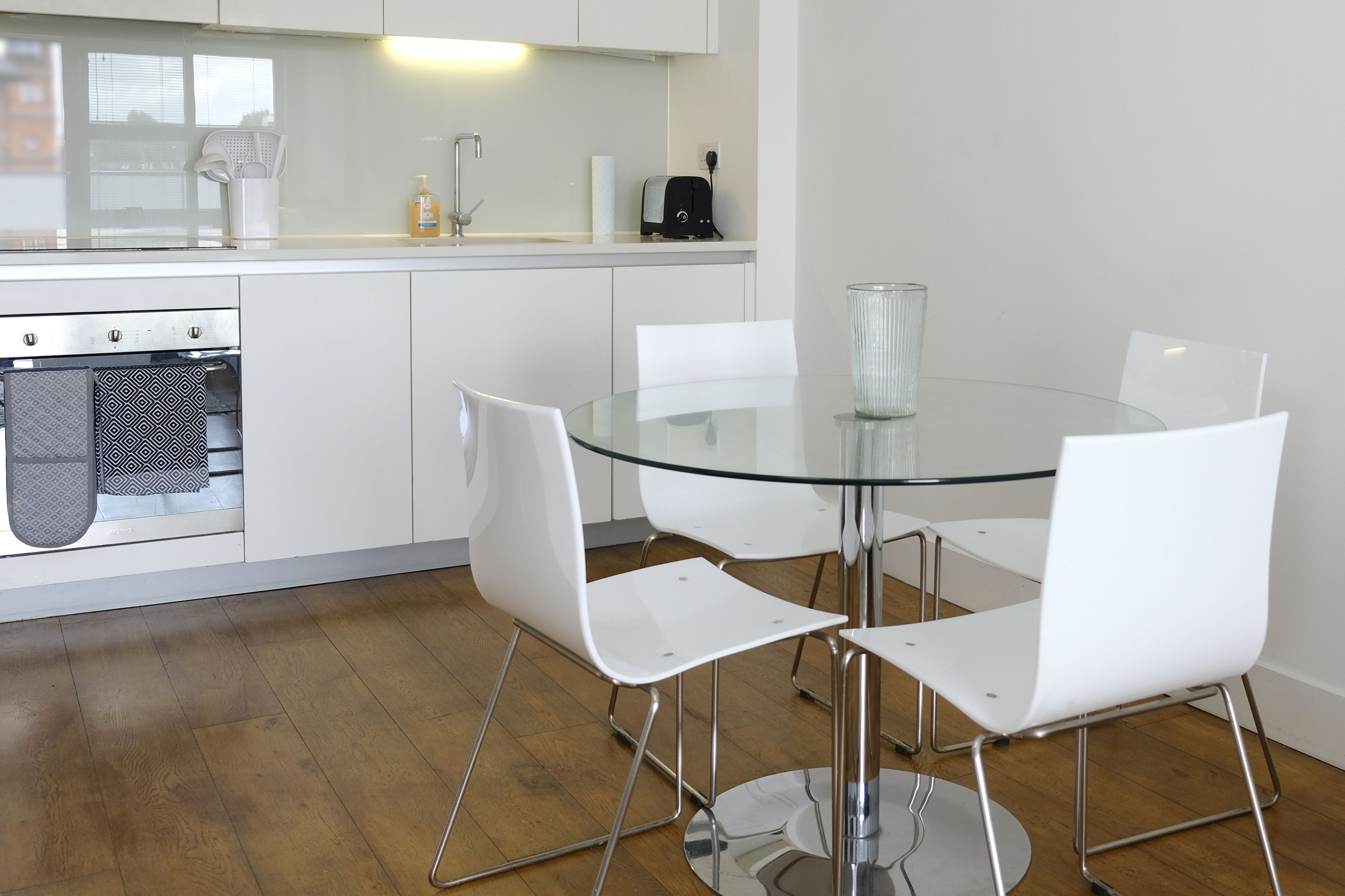 A modern kitchen with white cabinets, a glass dining table, and four white chairs.