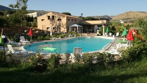 Una piscina con tumbonas y sombrillas, rodeada de vegetación y un edificio con tejado de baldosas.