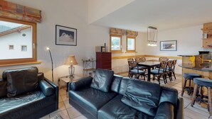 Ein Wohnzimmer mit schwarzem Ledersofa, eine Essecke mit Holztisch und Stühlen sowie eine Küchenzeile mit Bar.