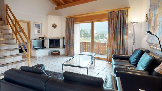 Ein Wohnzimmer mit Kamin, Fernseher, Sofa, Couchtisch und einem Balkon mit Vorhängen.
