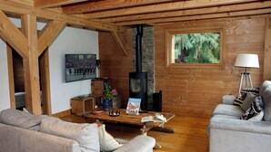 L’intérieur d’un chalet accueillant, avec un plafond en bois, une cheminée en pierre, un canapé, une table pour lire et une fenêtre donnant sur la forêt.
