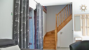Un salón con un sofá gris, un sillón negro, una escalera de madera y cortinas grises con estampado floral.