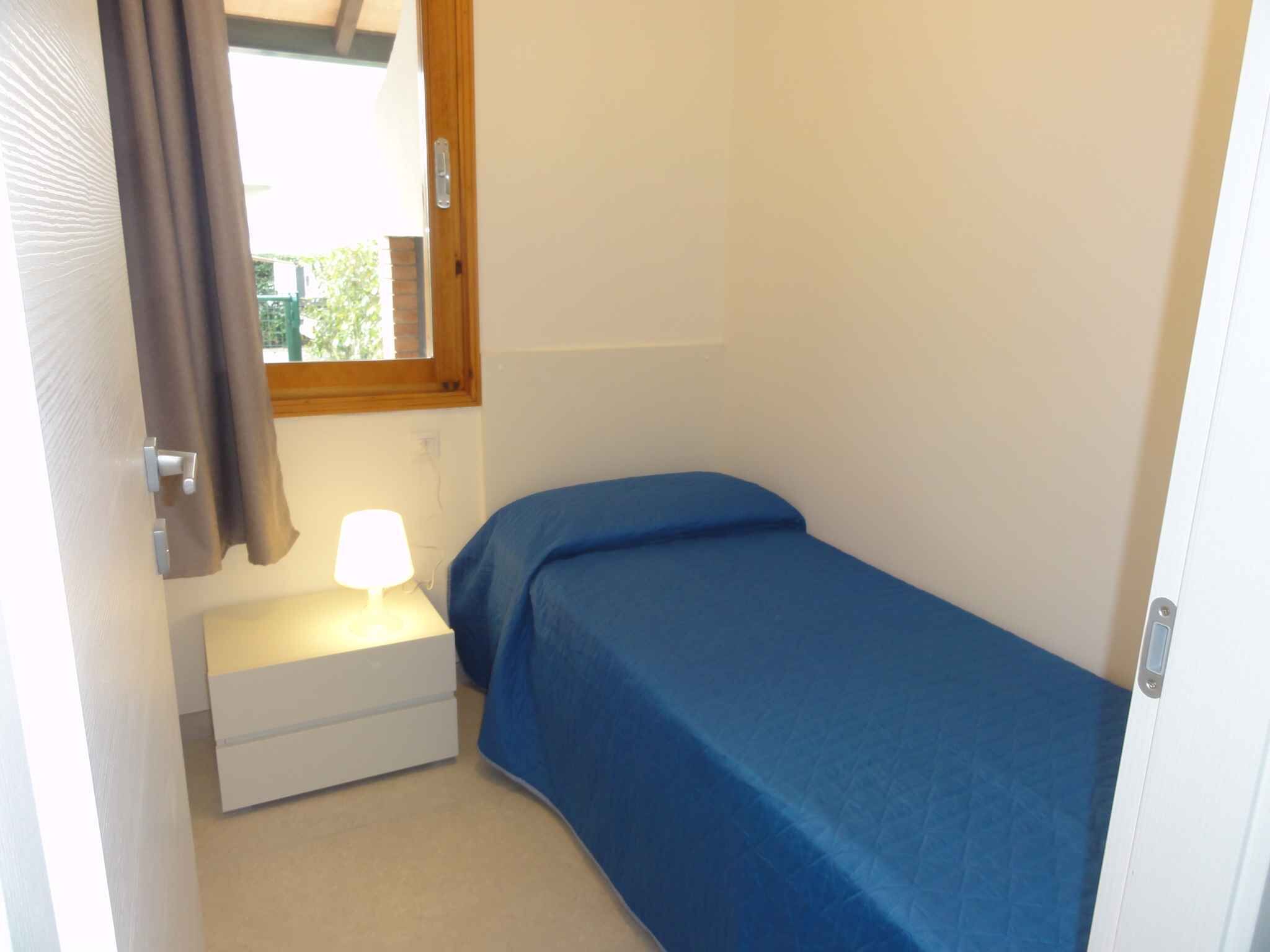 A small bedroom with a single bed, a nightstand with a lamp, a window with curtains, and a door.