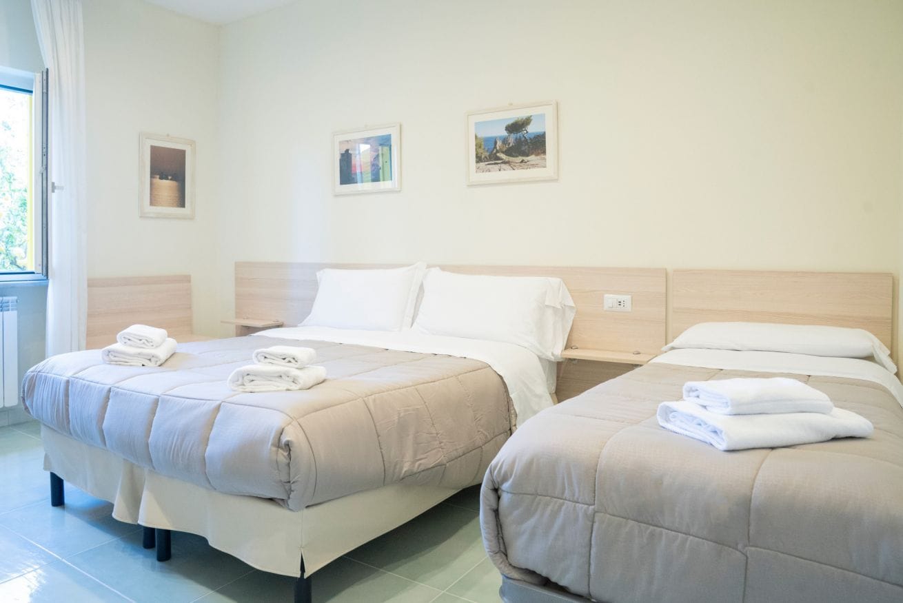 Ein Hotelzimmer mit zwei Betten, jeweils mit einer beige farbenen Bettdecke und weißen Kissen. An der Wand hängen gerahmte Bilder, und das Zimmer hat ein Fenster mit Vorhängen.