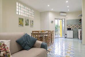 A living room with a sofa, a dining table with chairs, a kitchen area with cabinets, and a tiled floor.