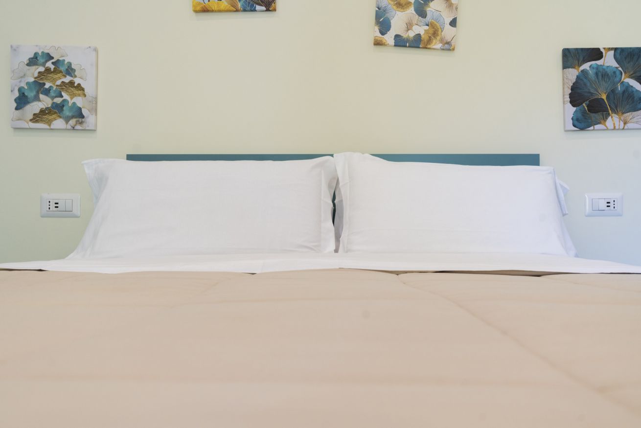 Una cama con sábanas blancas y un cabecero verde azulado, con dos tomas de corriente en la pared.