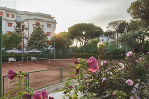 Una cancha de tenis con una red, rodeada por una valla. Se ven árboles y un edificio con balcones al fondo.