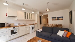A modern kitchen with white cabinets, a built-in oven, and a washing machine. There is a grey sofa with orange and white cushions, and a wooden coffee table. - Pauillac two bedroom apartment on second floor (Blaye)