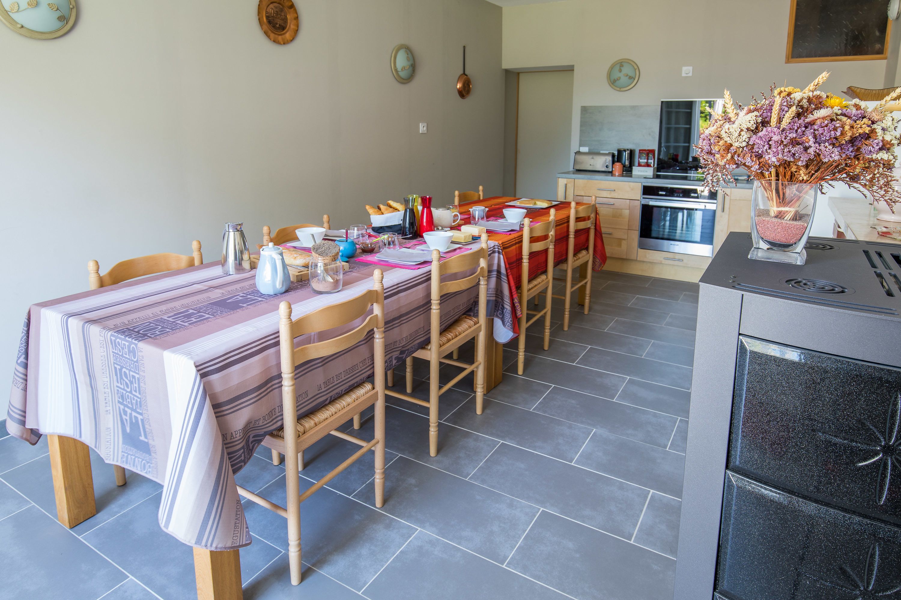 Comedor con mesa preparada para cuatro, sillas de madera, cocina al fondo y un florero con flores en la encimera.