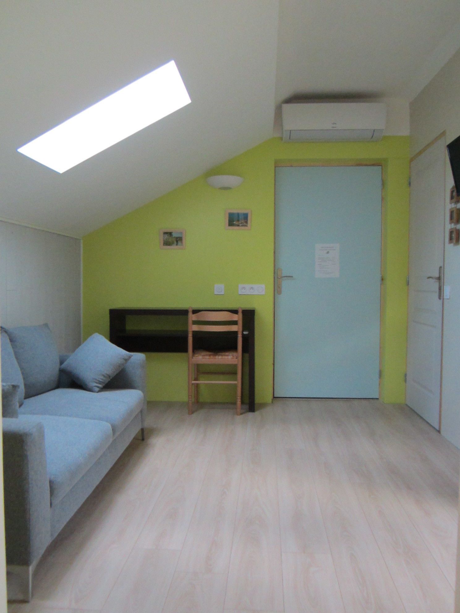 Una habitación con techo inclinado, una cama, una silla, una puerta y un aire acondicionado instalado en la pared.