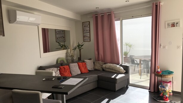 A modern living room with a grey sofa, a dining table, and a balcony with a view. - RENTED VILLA F3 RENTED IN THE GREEN AREA (Sainte-Maxime)