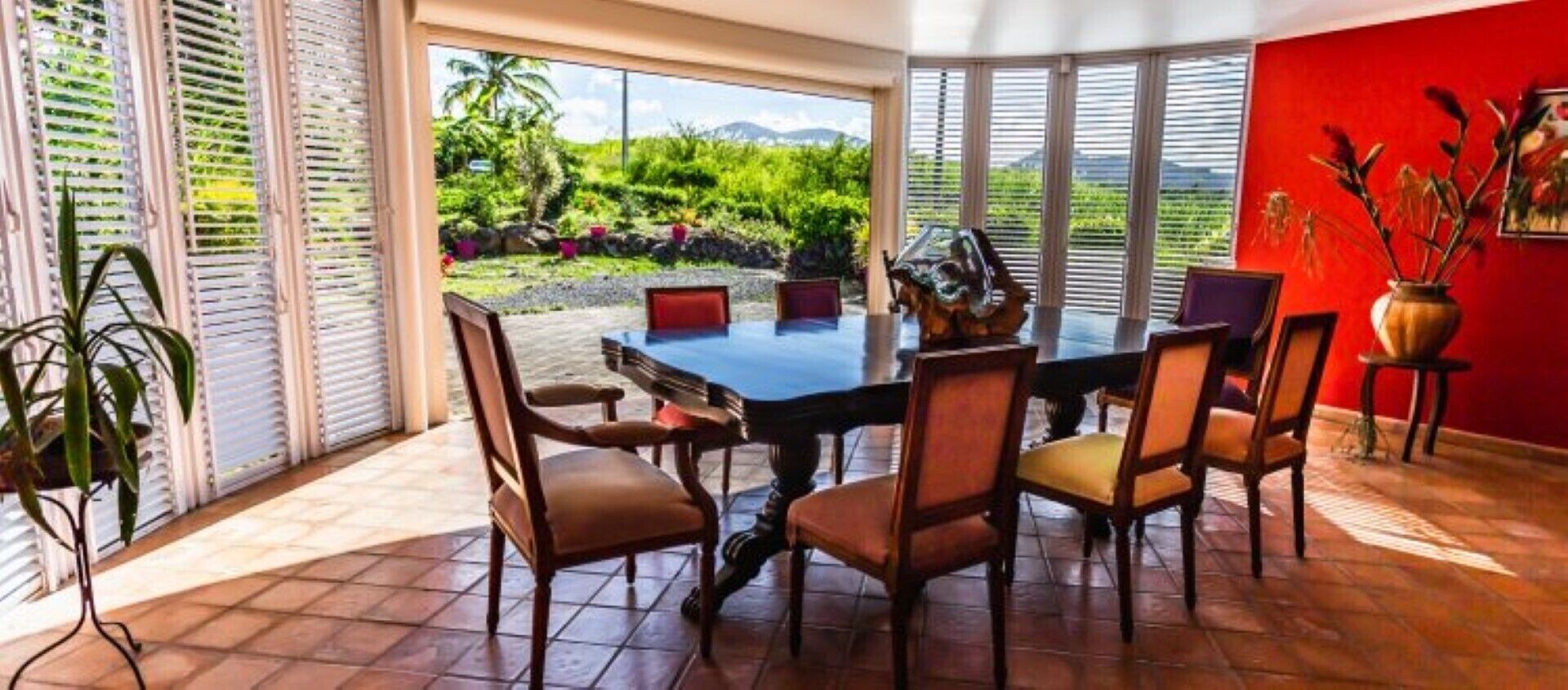 A dining area with a large table and chairs, large windows with blinds, a red wall, and a view of greenery outside.