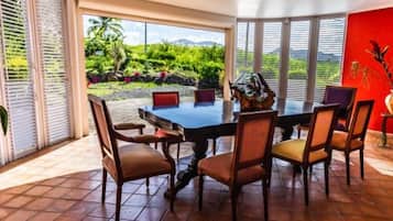 Un espace repas avec une grande table et des chaises, de grandes fenêtres équipées de stores, un mur rouge et une vue sur la verdure à l’extérieur.