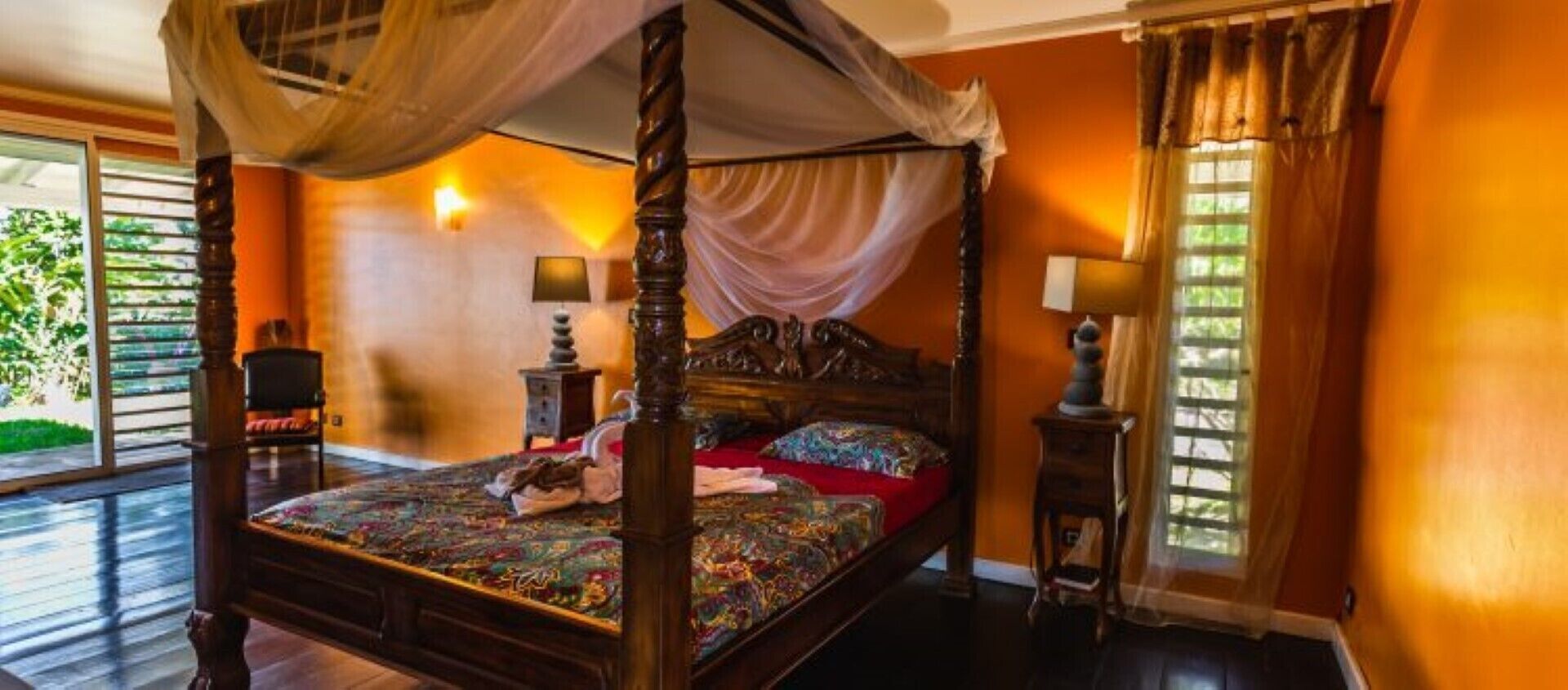 A four-poster canopy bed with a colorful bedspread, a nightstand with a lamp, and a window with curtains.
