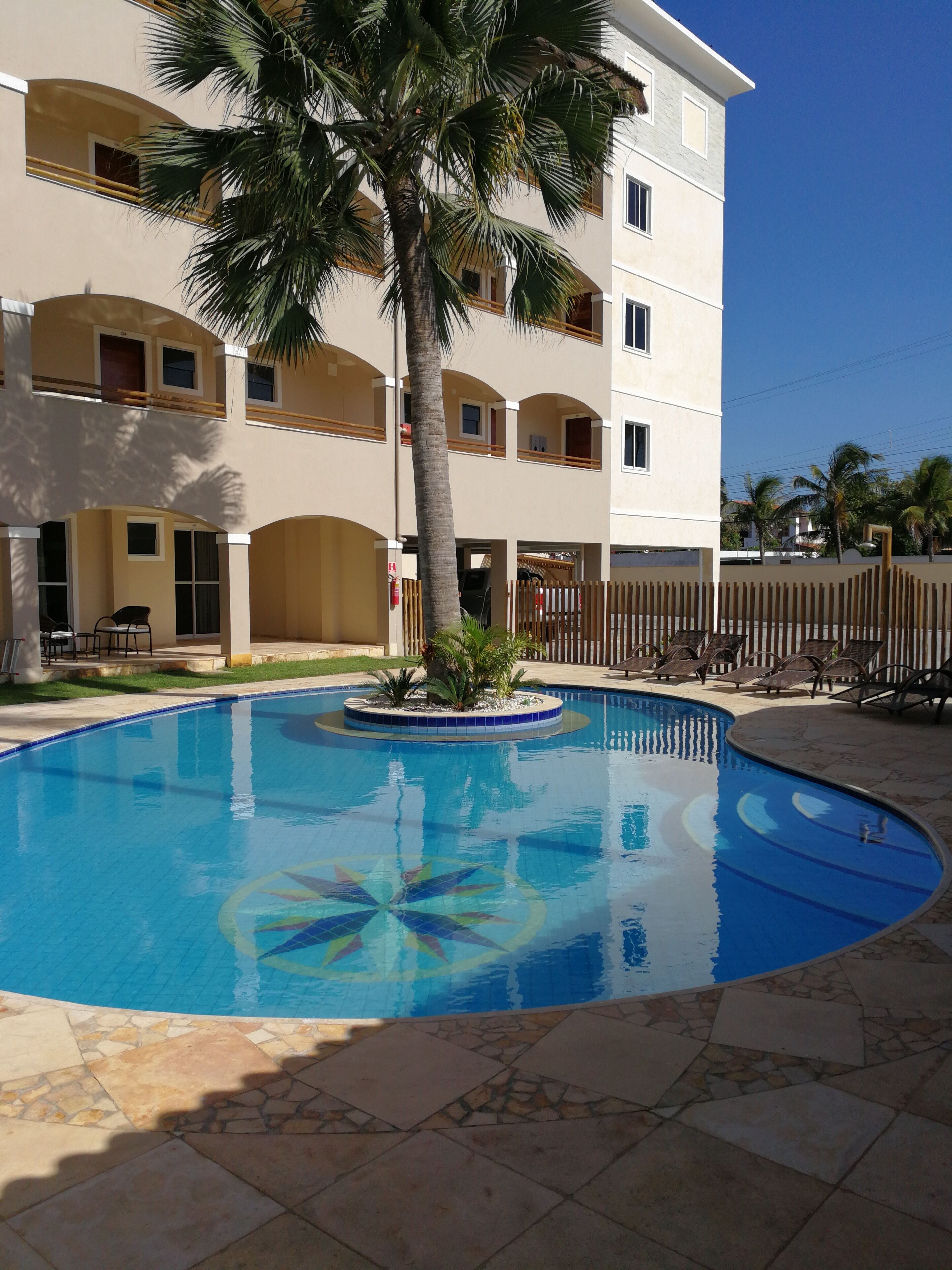 Ein Swimmingpool mit einer Palme, einer überdachten Terrasse und einem mehrstöckigen Gebäude mit Balkonen.