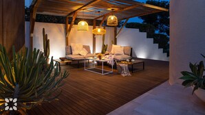 Een terras buiten met houten vloer, twee comfortabele banken, een bijzettafeltje en hangende lampen.