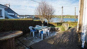 Un patio al aire libre con una mesa y sillas de madera, un camino de piedra y vistas a un cuerpo de agua y edificios al fondo.