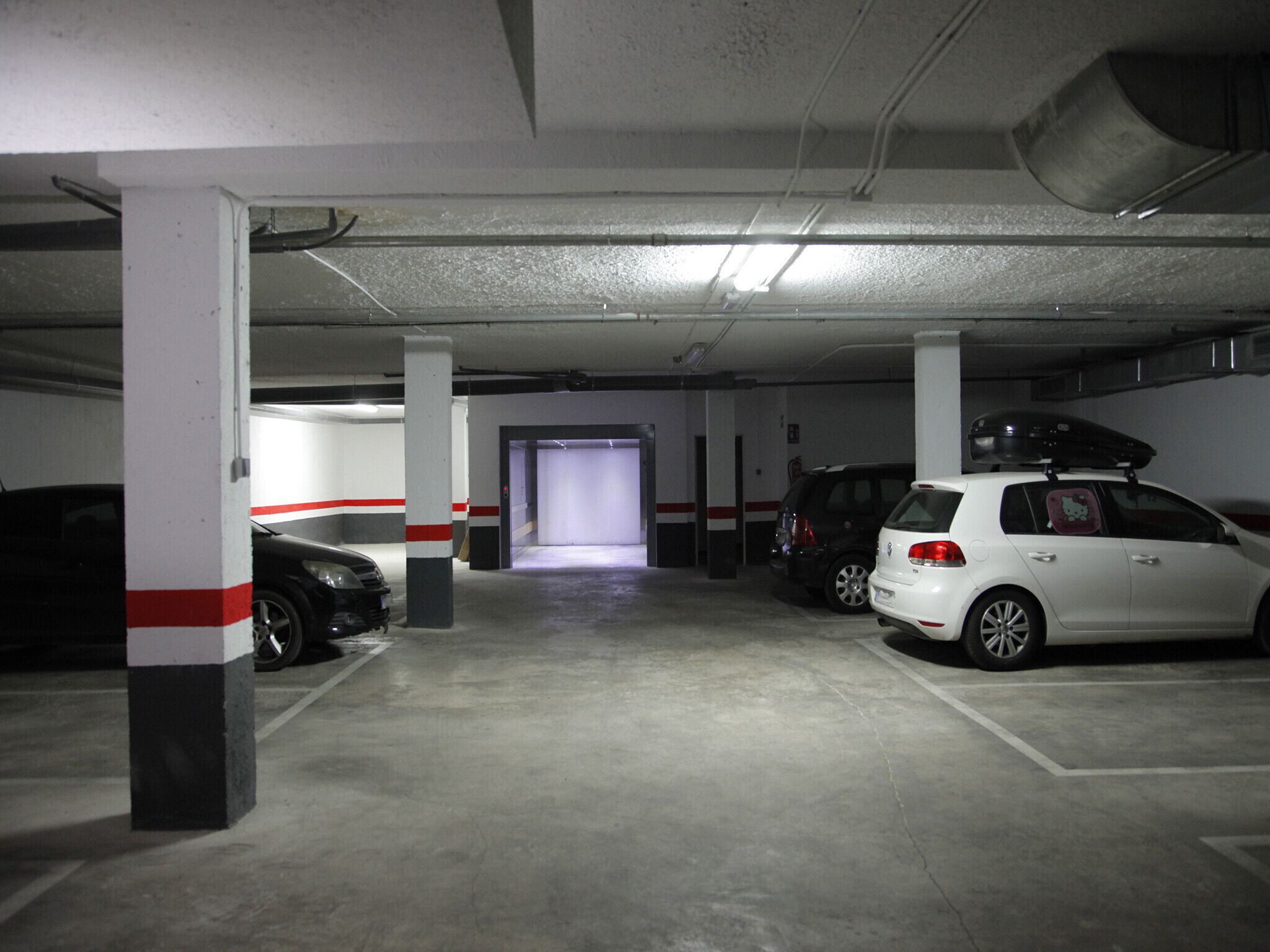 A parking garage with concrete pillars, a white car with a Hello Kitty decal, and a black car parked in the distance.