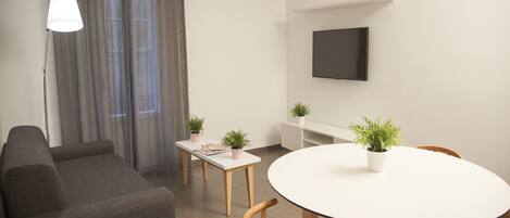Una sala moderna con un sofá gris, una mesa de comedor redonda con sillas de madera, una mesita auxiliar blanca y dos plantas en macetas.