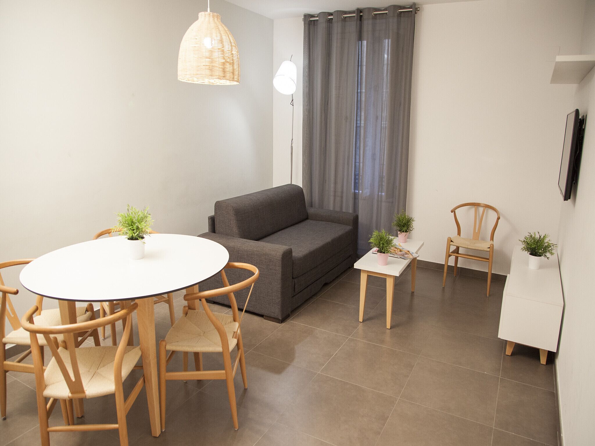 A modern living room with a grey sofa, a round dining table with chairs, a small side table, and two potted plants.