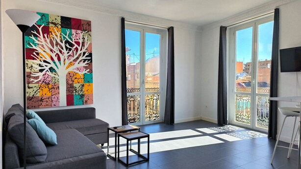 A modern living room with a gray sofa, a coffee table, and a colorful abstract painting. Large windows with black curtains provide a view of the cityscape. - Flats Friends Torres Quart 1 bedroom with terrace (Valencia)