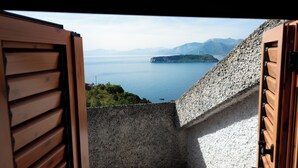 Une vue depuis un balcon, avec un mur en pierre et des volets en bois, donnant sur une étendue d’eau et des montagnes lointaines.