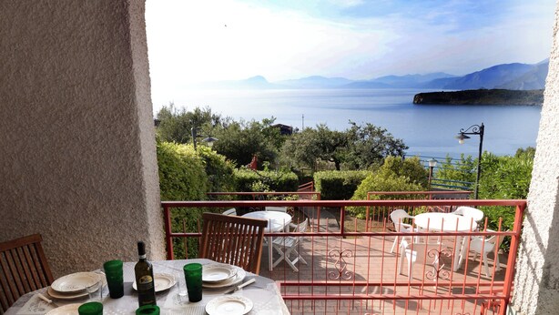 Une table à manger dressée avec des assiettes, des verres et des couverts, sur un balcon offrant une vue panoramique sur la mer et les montagnes.
