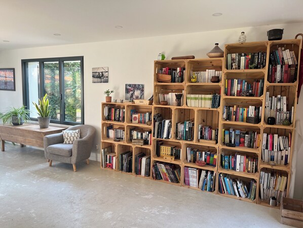 Uma sala de estar espaçosa com uma grande estante de madeira cheia de livros, uma poltrona cinza confortável, uma mesa de centro de madeira e um vaso com planta.