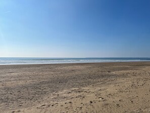 Uma extensa praia de areia, com pegadas e céu claro.