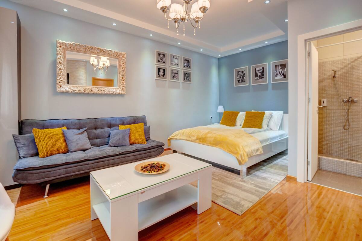 A modern bedroom with a grey sofa, a white bed, and a small table with a bowl of fruit.