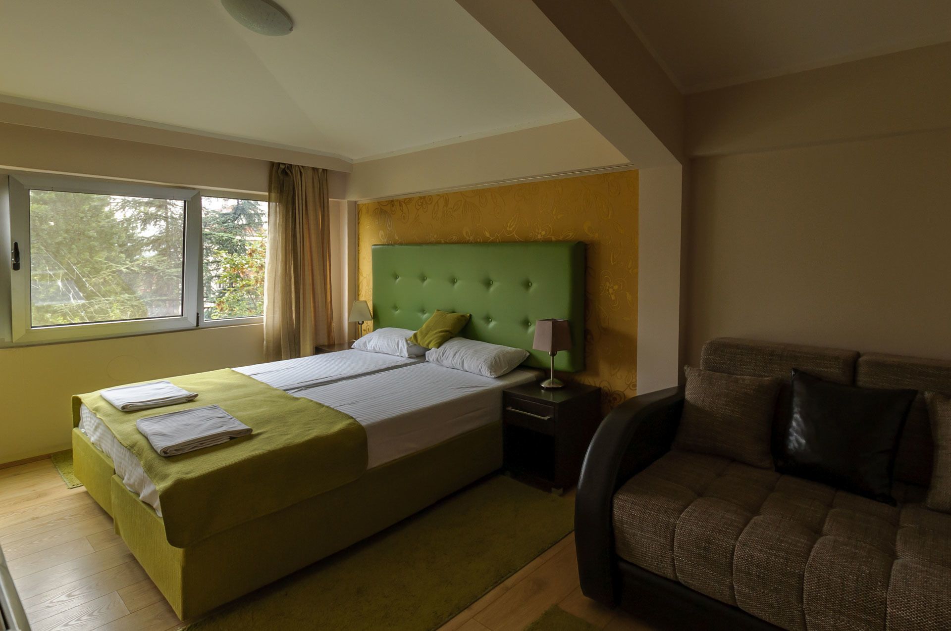 A hotel room with a large bed, two bedside tables, a green headboard, a window with curtains, and a brown sofa.