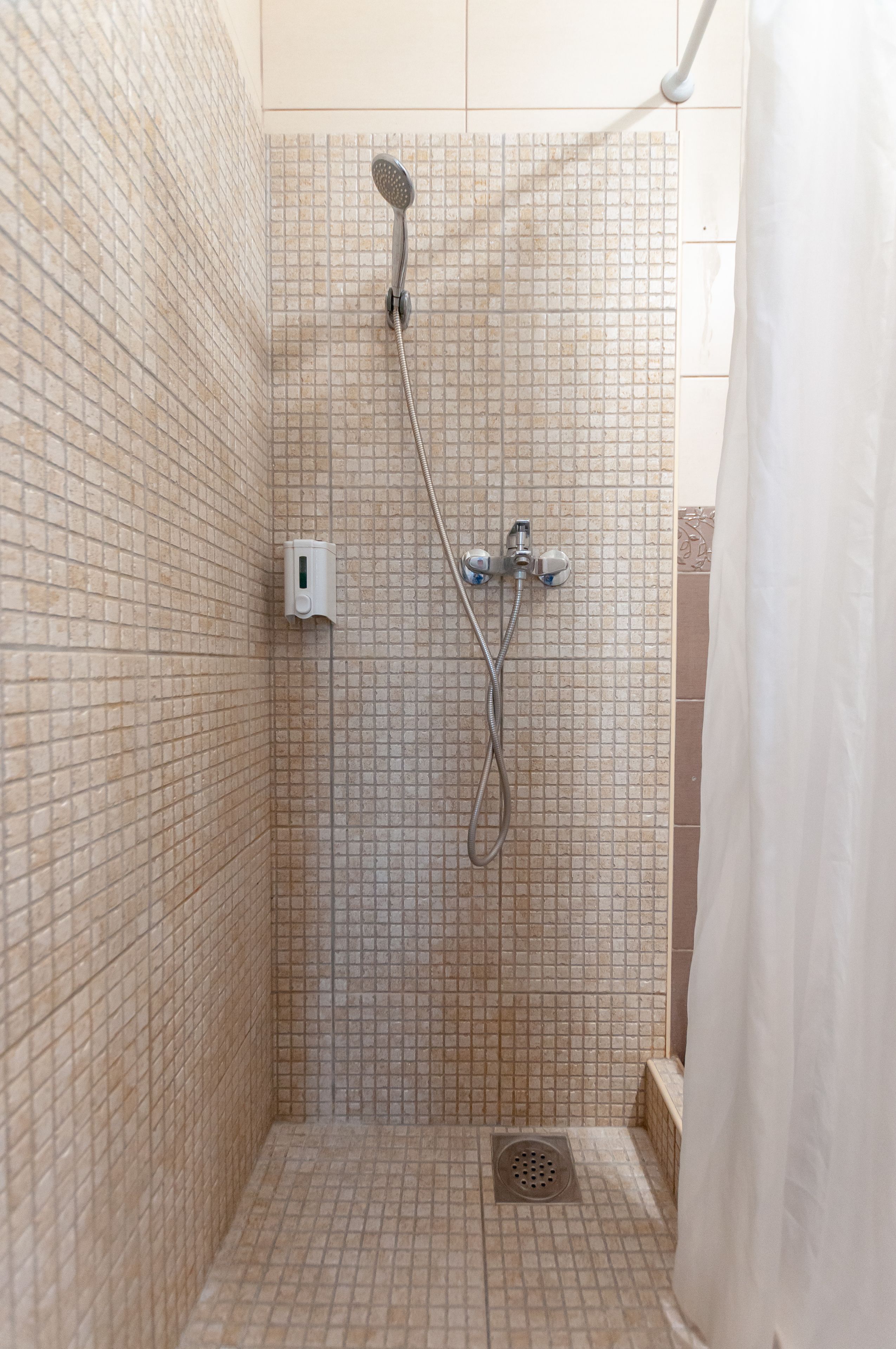 Área de duche com parede revestida em azulejos, chuveiro manual, torneira fixa na parede e ralo.