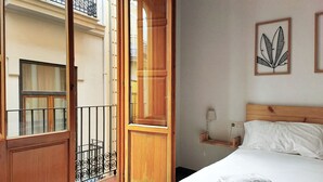 A hotel room with a bed, a nightstand, a wall-mounted lamp, and a balcony with a view of the building's exterior.