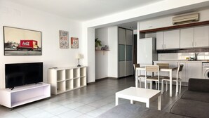 A modern living room with a flat-screen TV, a white console table, a grey sofa, and a dining area with a wooden table and chairs.