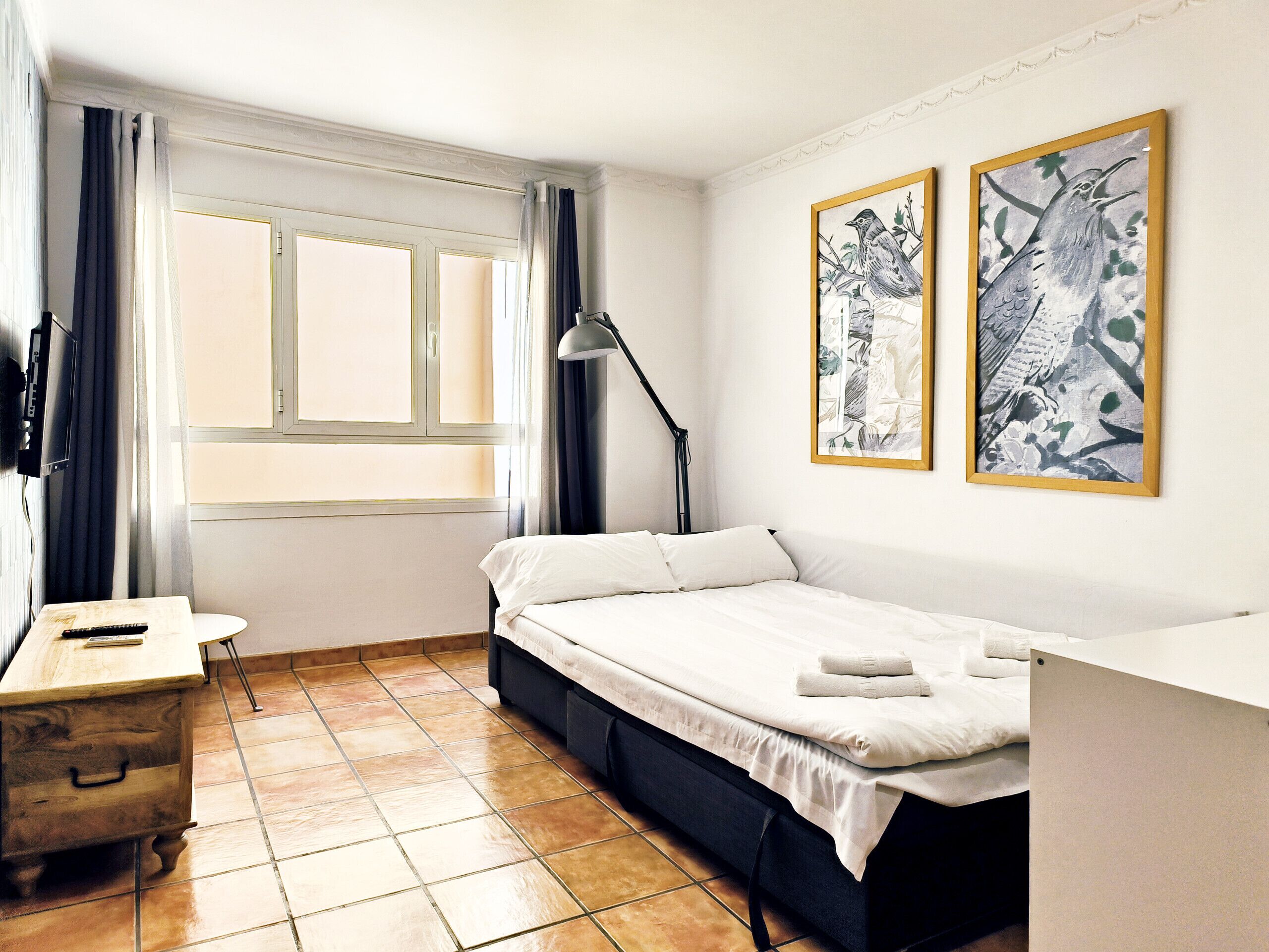 A bedroom with a bed, a desk, a chair, a TV, and two framed artworks on the wall.
