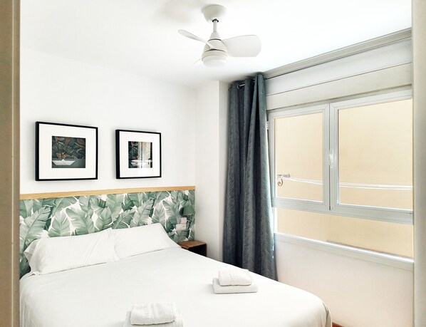 A hotel room with a bed, two framed pictures on the wall, a ceiling fan, a window with curtains, and a view of the outdoors.