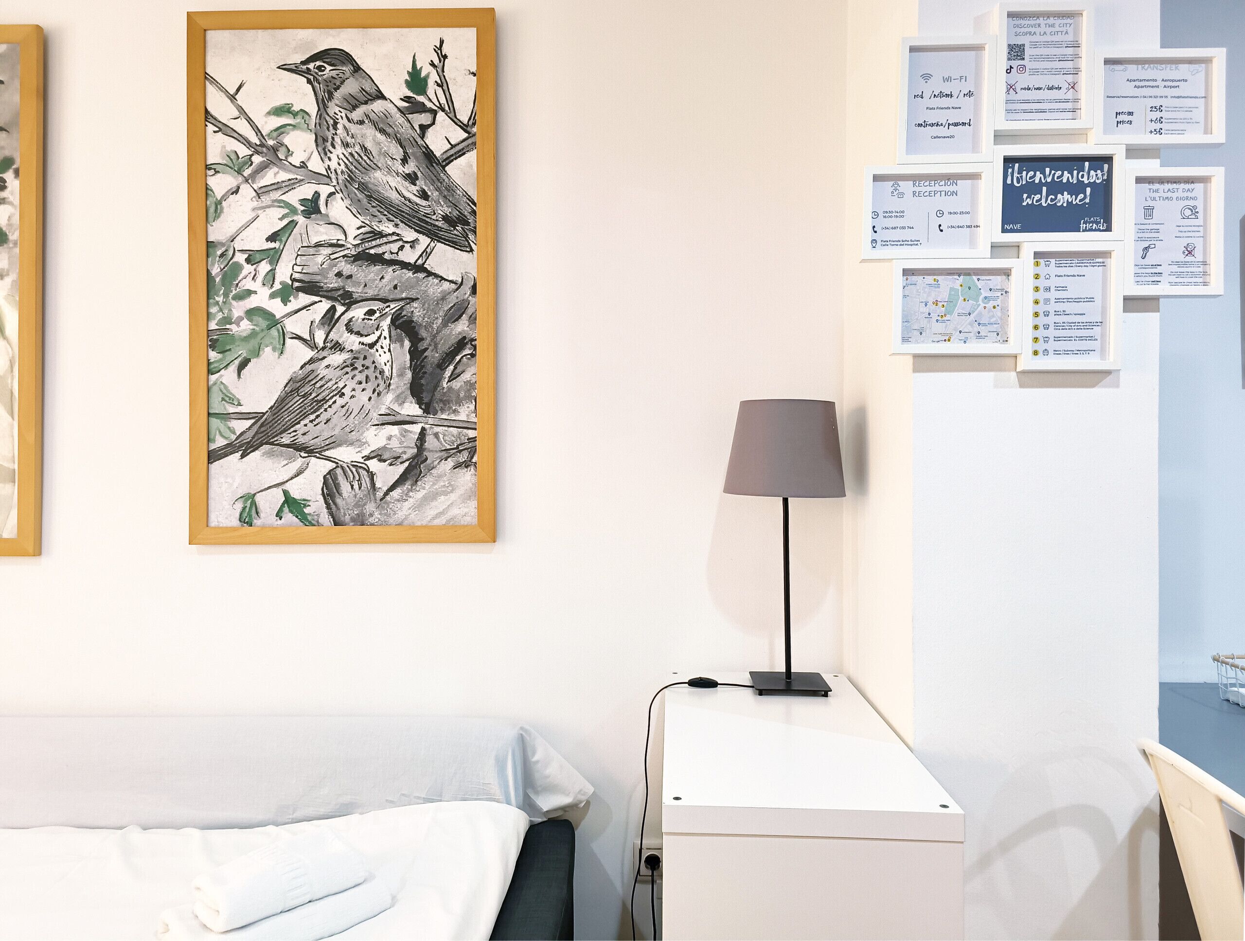 Habitación de hotel con cama, escritorio, lámpara y un muro con varios carteles informativos.