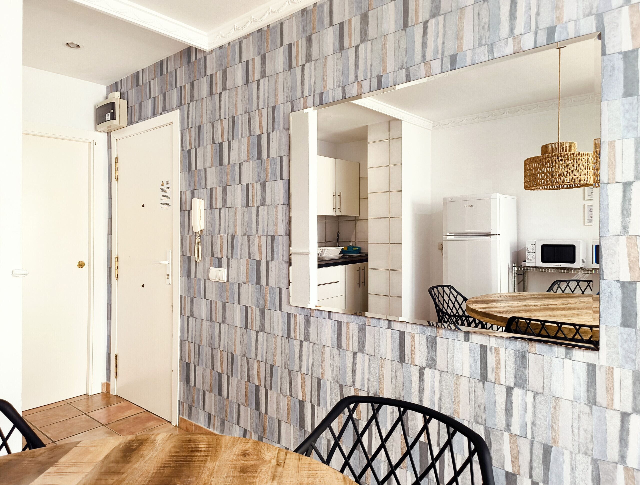 A dining area with a wooden table and chairs, a large mirror reflecting the kitchen, and a patterned wall.