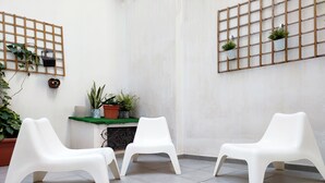 A room with three white plastic chairs arranged in a semi-circle, a small table with potted plants, and a wall-mounted shelf with plants and a decorative item.
