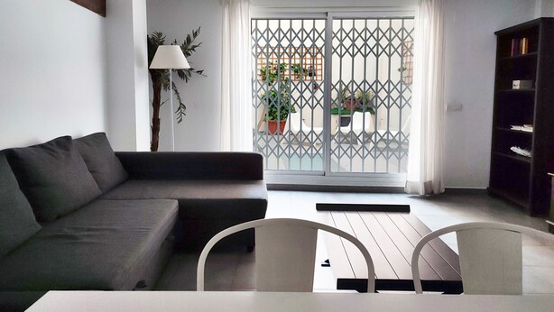 A modern living room with a sofa, a dining table, and chairs. There is a sliding glass door leading to an outdoor area with plants and a chair.
