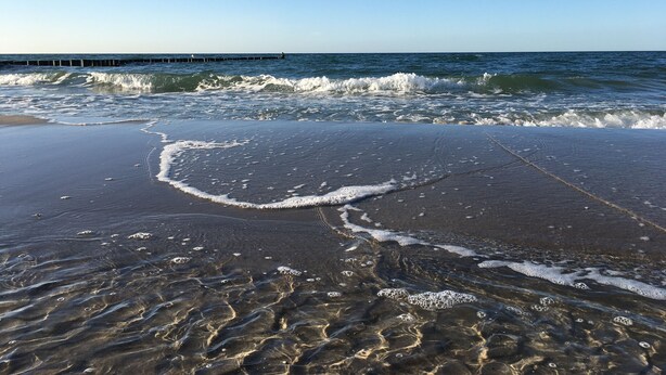 A beach with clear water, waves, and a pier in the background.