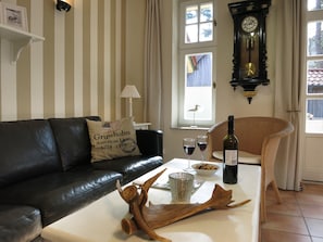 Uma sala de estar com parede listrada, um sofá de couro preto, uma mesa branca com uma garrafa de vinho, copos e um objeto decorativo de madeira.